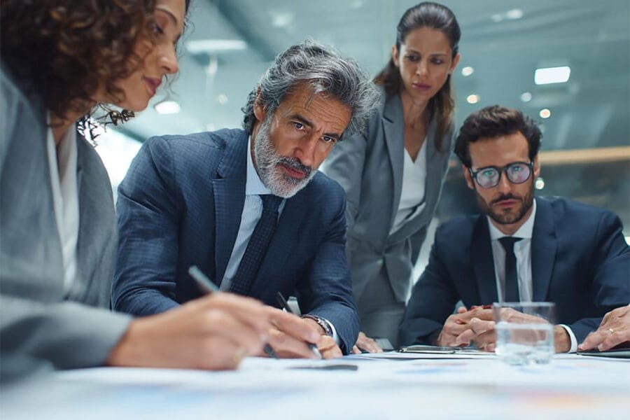 vendor-sprawl - Marinum Consulting Group of four professionals in a modern office, one man signing documents while others observe at a table with papers and water.